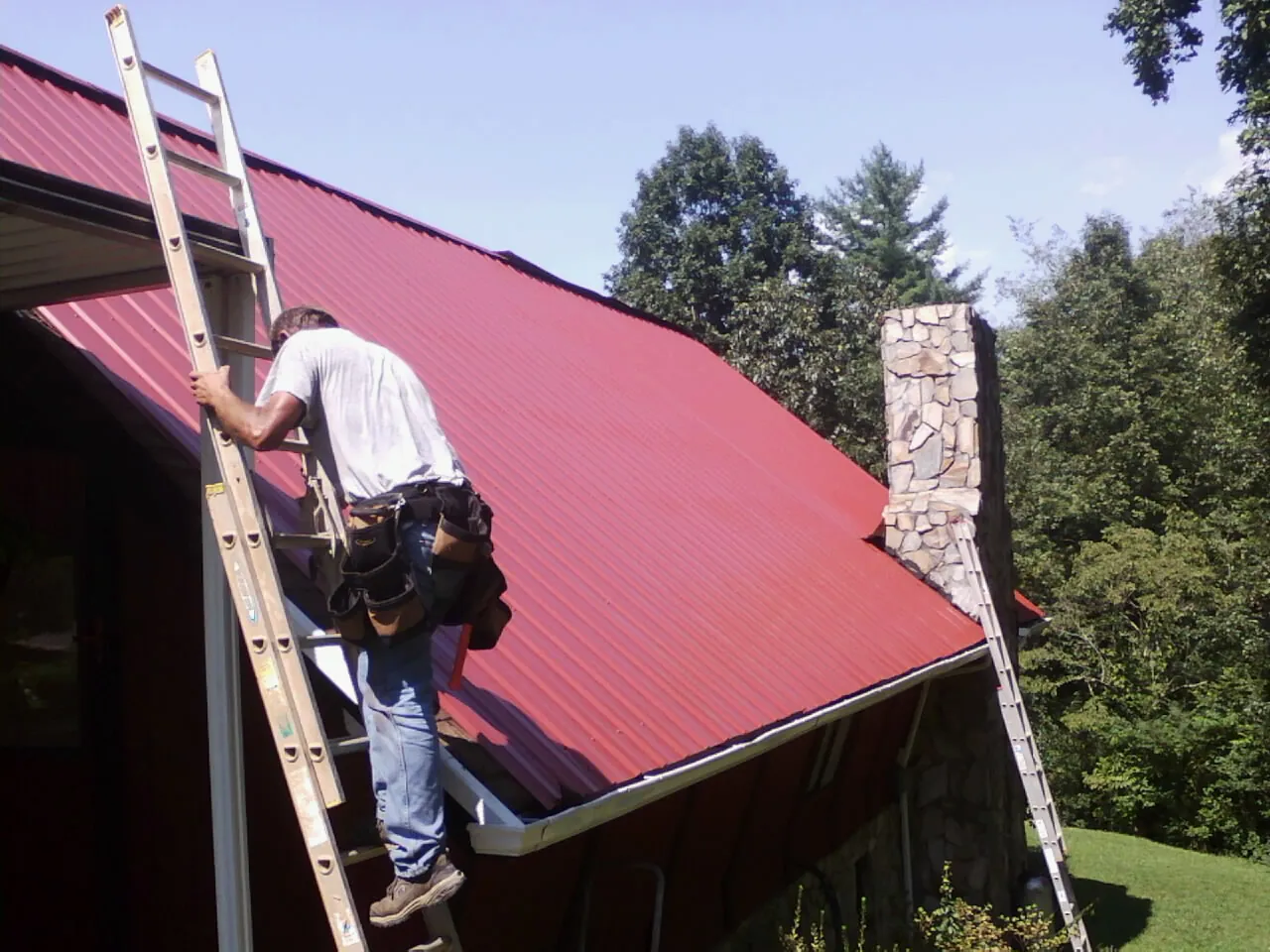 Roofer on a ladder beside a red metal roof and stone chimney — Soffit & Fascia Repair by Roofing Brothers in Chesapeake City